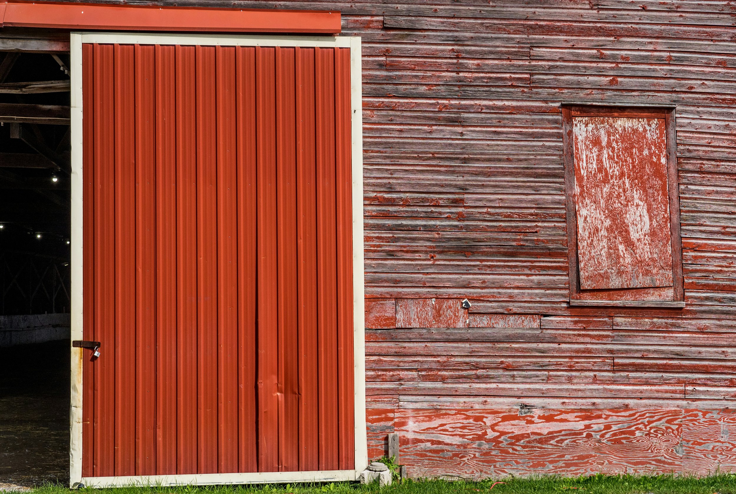 Red barn exterior with peeling paint, a closed red sliding door, and a small boarded-up window.
