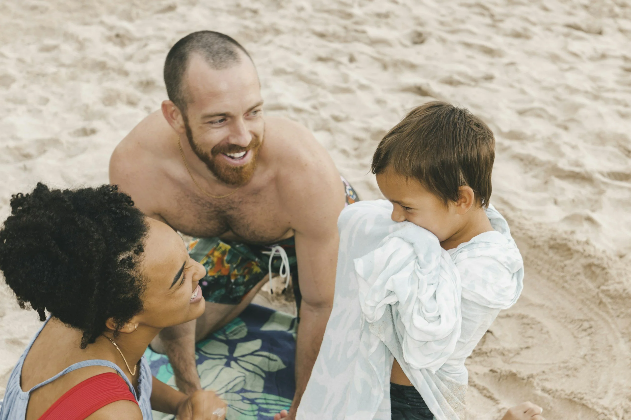 A smiling woman with dark curly hair, a shirtless man with a beard, and a young boy with brown hair on a sandy beach. The boy is playfully covering his face with a light-colored hoodie while the woman looks at him.