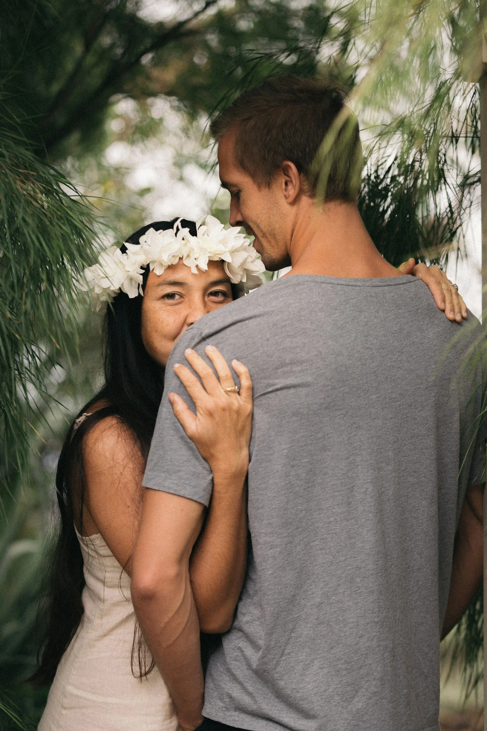 A couple embracing outdoors, woman with a floral crown looking at the camera, man with back to camera, surrounded by green foliage.