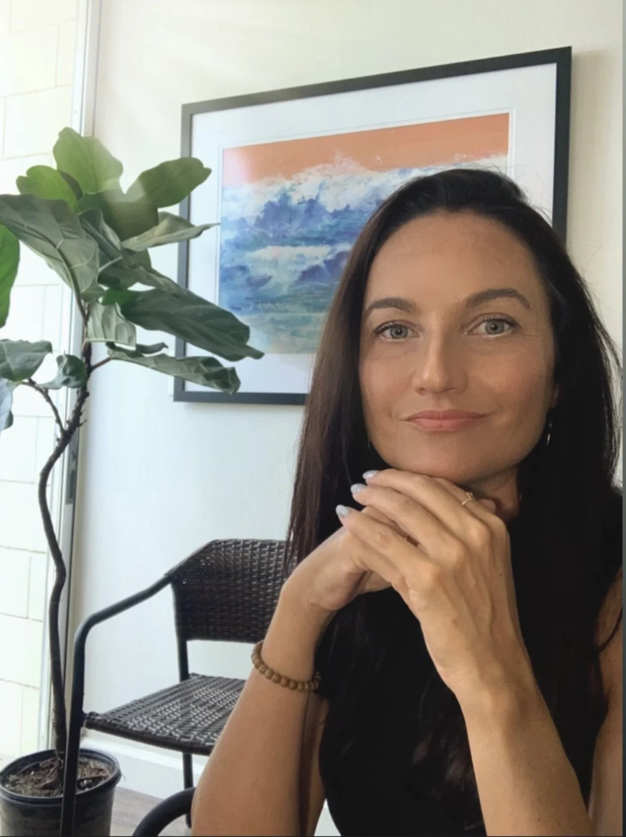 A woman with long dark hair and pierced ears poses with her hands under her chin, smiling slightly. She is indoors, next to a large green plant and a framed colorful abstract painting on the wall.