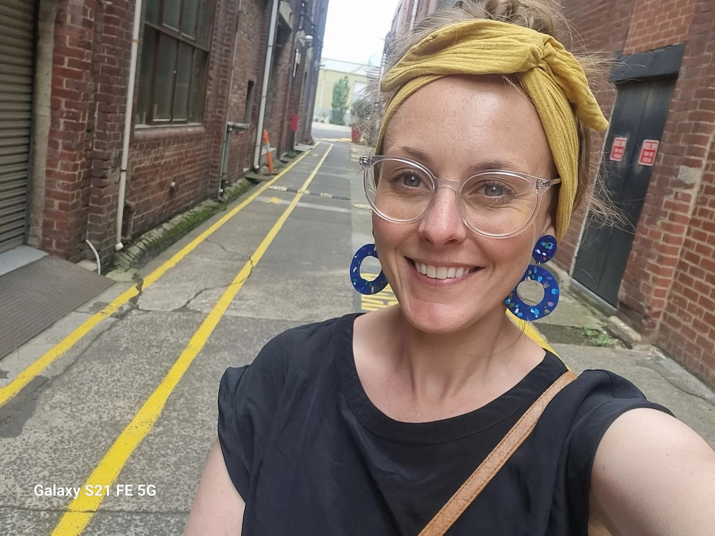 Kristin Sargent — Trauma-Informed Creative Coach, Mentor & Somatic Practitioner — smiling in a bright urban laneway, wearing clear glasses, a yellow headscarf and blue statement earrings