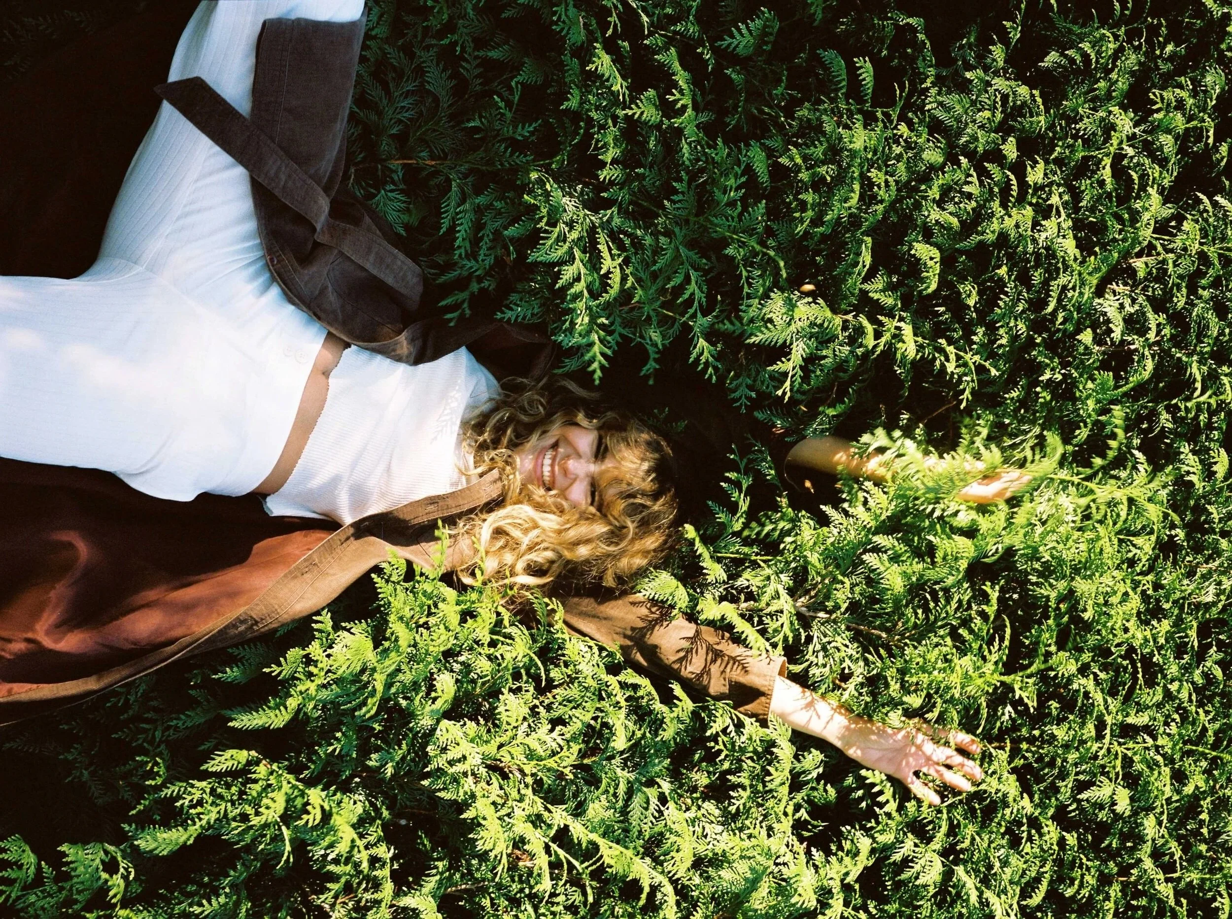 A woman with curly hair lying on her back among green ferns, smiling with one arm extended outward.