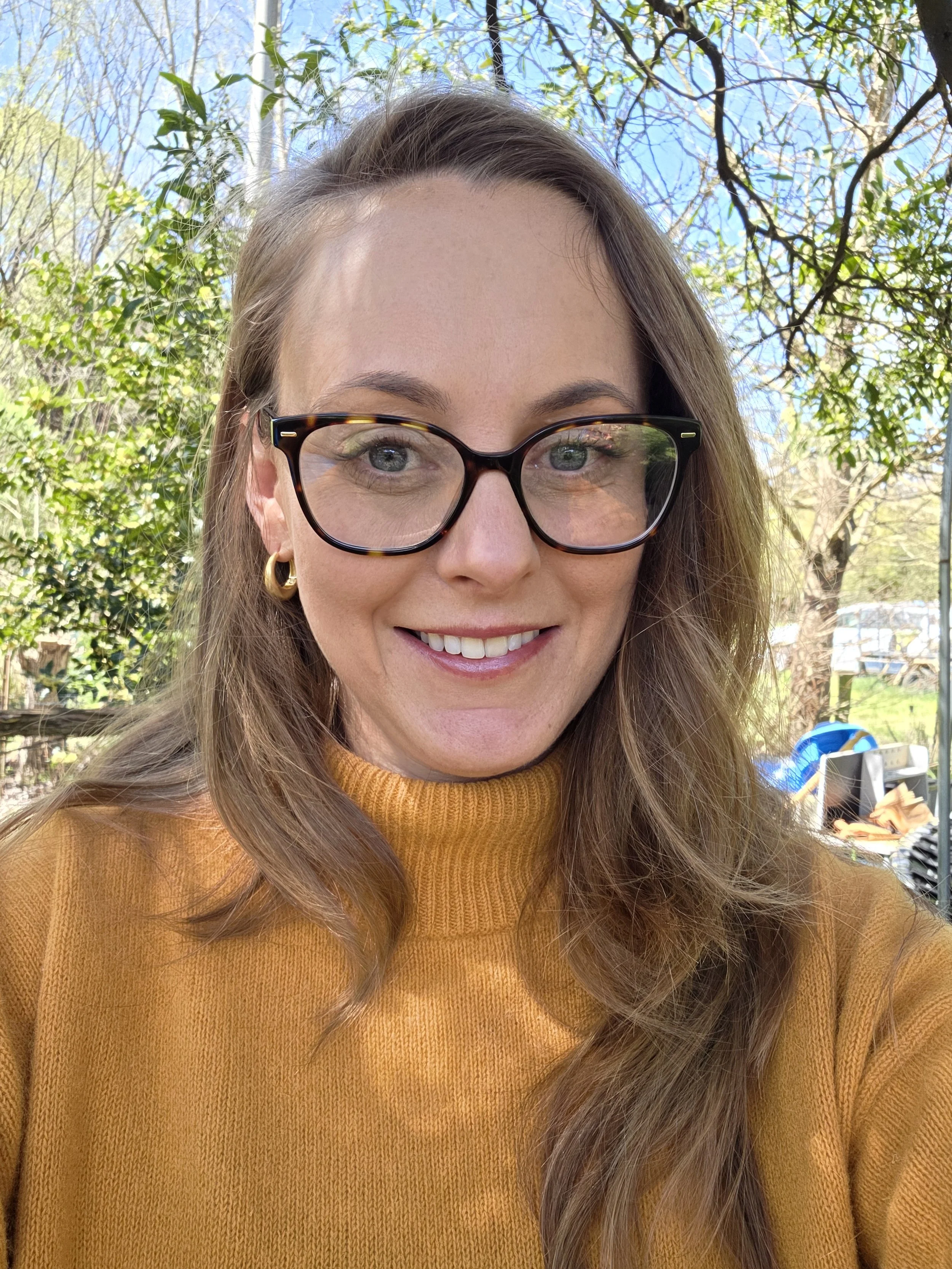 A woman with long brown hair wearing large black glasses and a mustard yellow turtleneck sweater. She is smiling and outdoors with trees, a blue sky, and a backyard visible in the background.