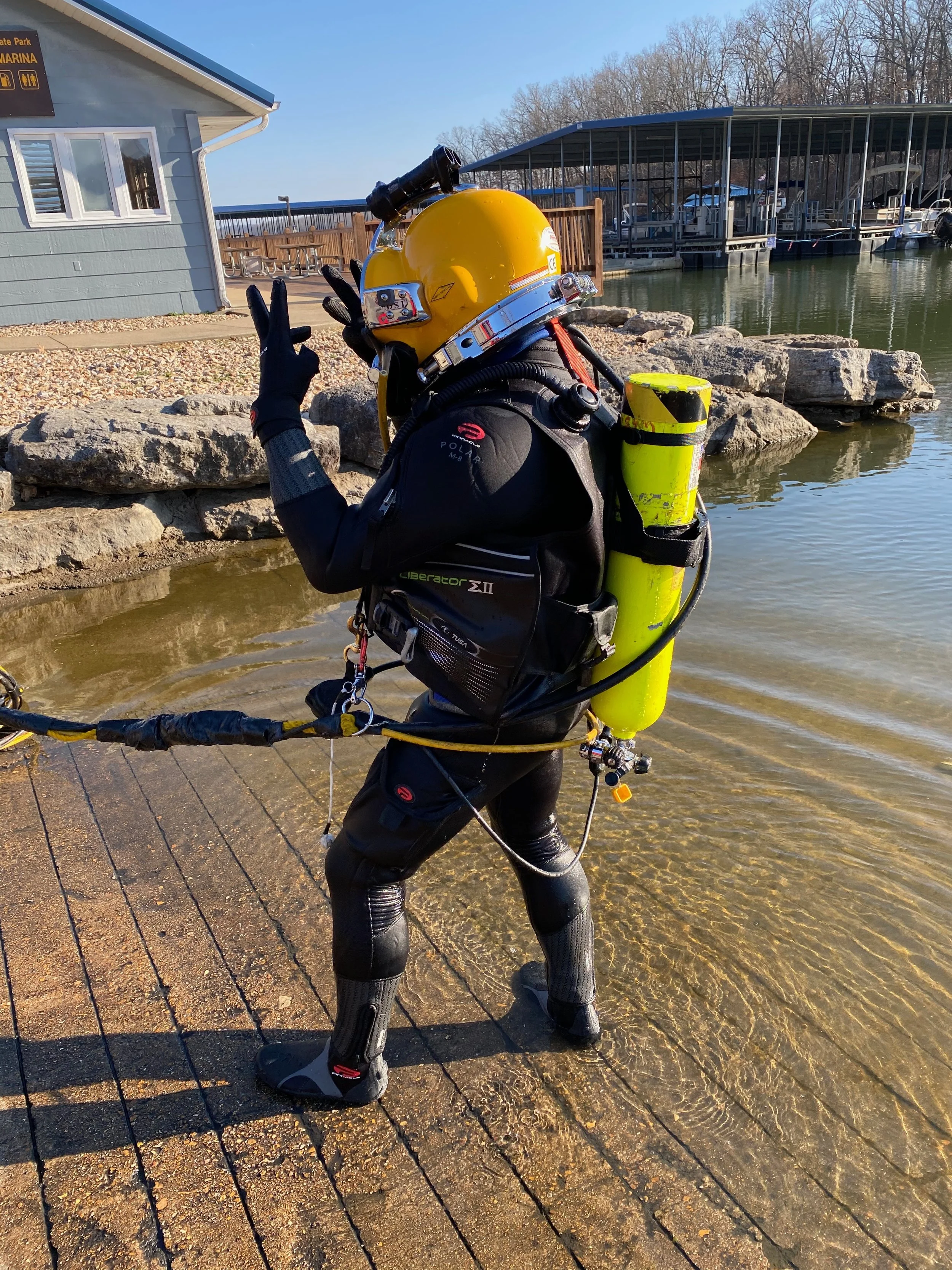 A diver in a wetsuit and scuba gear standing on a wooden dock by the water, holding a yellow helmet and making a peace sign with their hand.