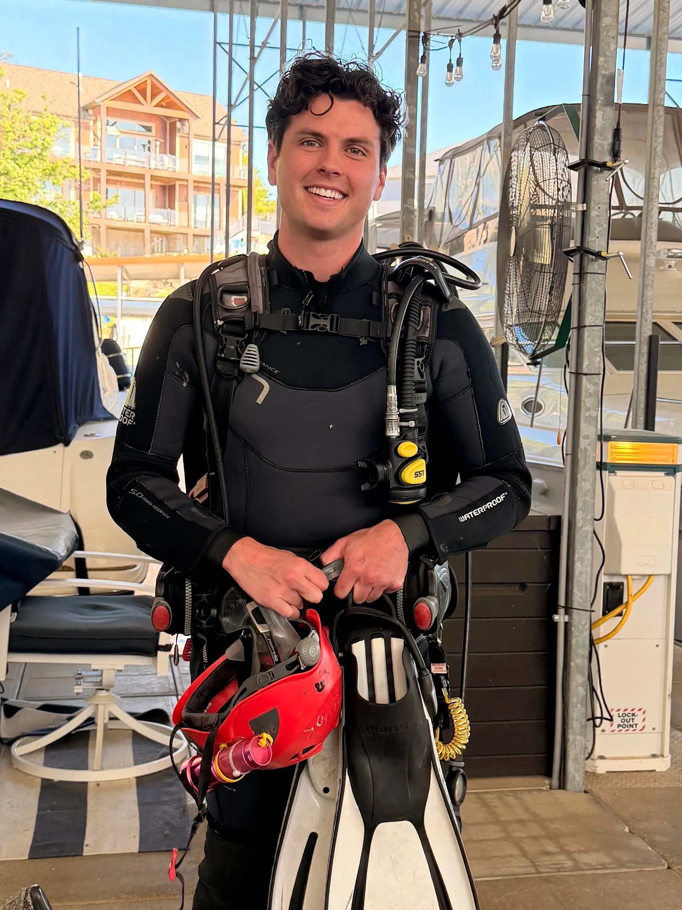 A young man smiling in scuba gear, standing outdoors under a tent with buildings and trees in the background.