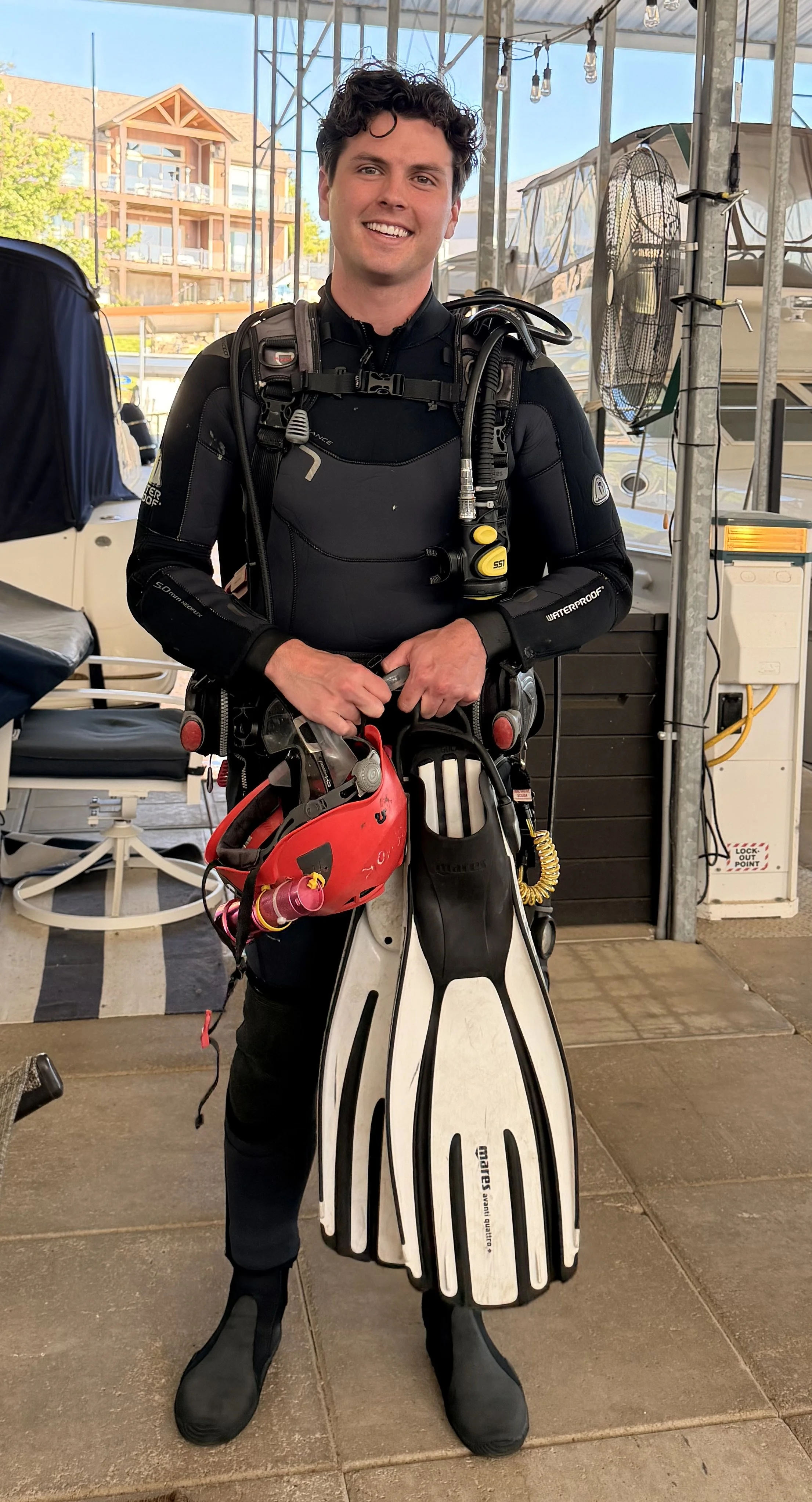 A young man dressed in diving gear smiling, holding a red diving helmet and a pair of diving fins, standing indoors with a boat and buildings visible in the background.