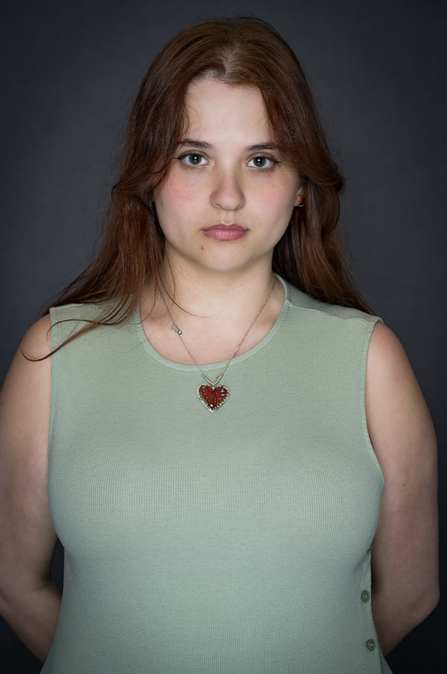 Chesney is wearing a green tank top, a heart necklace, and wearing her auburn hair down in this headshot.