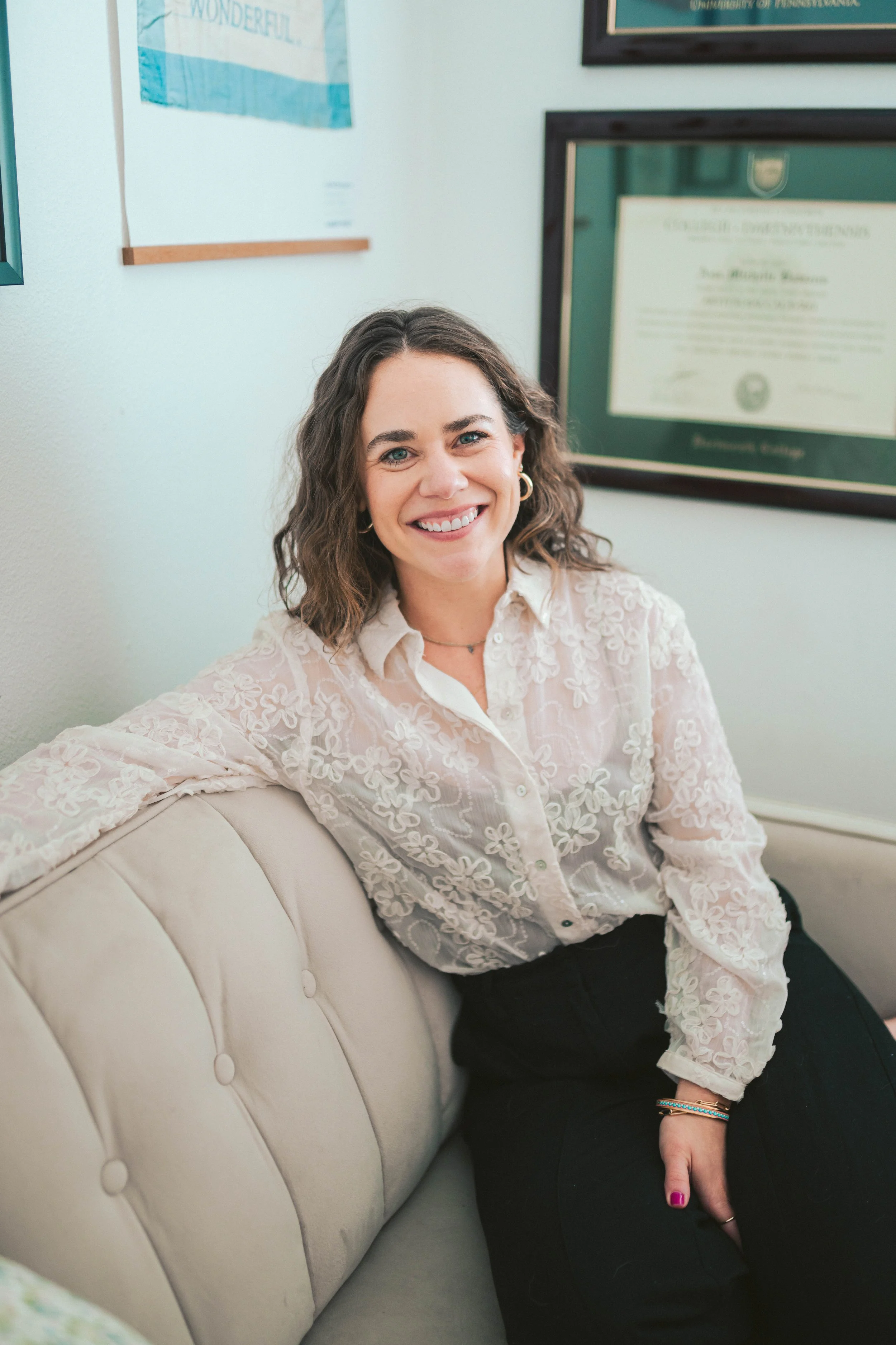 A smiling woman with curly brown hair, wearing a cream-colored lace blouse, seated on a beige sofa in an office. Behind her are framed diplomas and certificates on the wall.