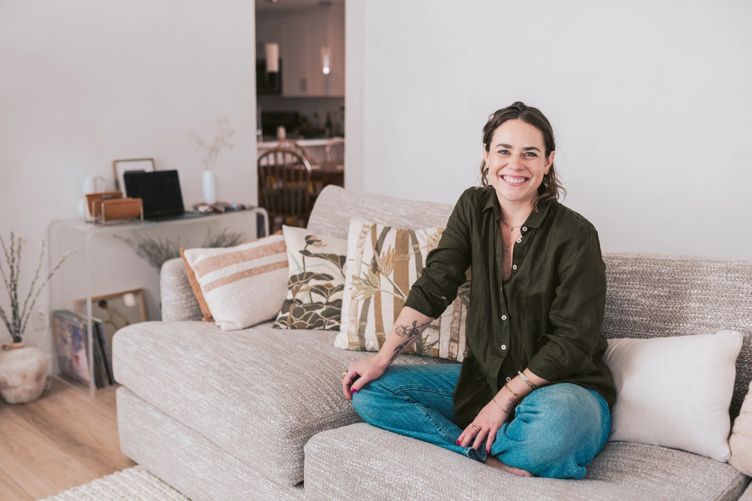 A woman with dark hair and tattoos sitting cross-legged on a beige sofa, smiling in a living room with decorative pillows and a white wall.