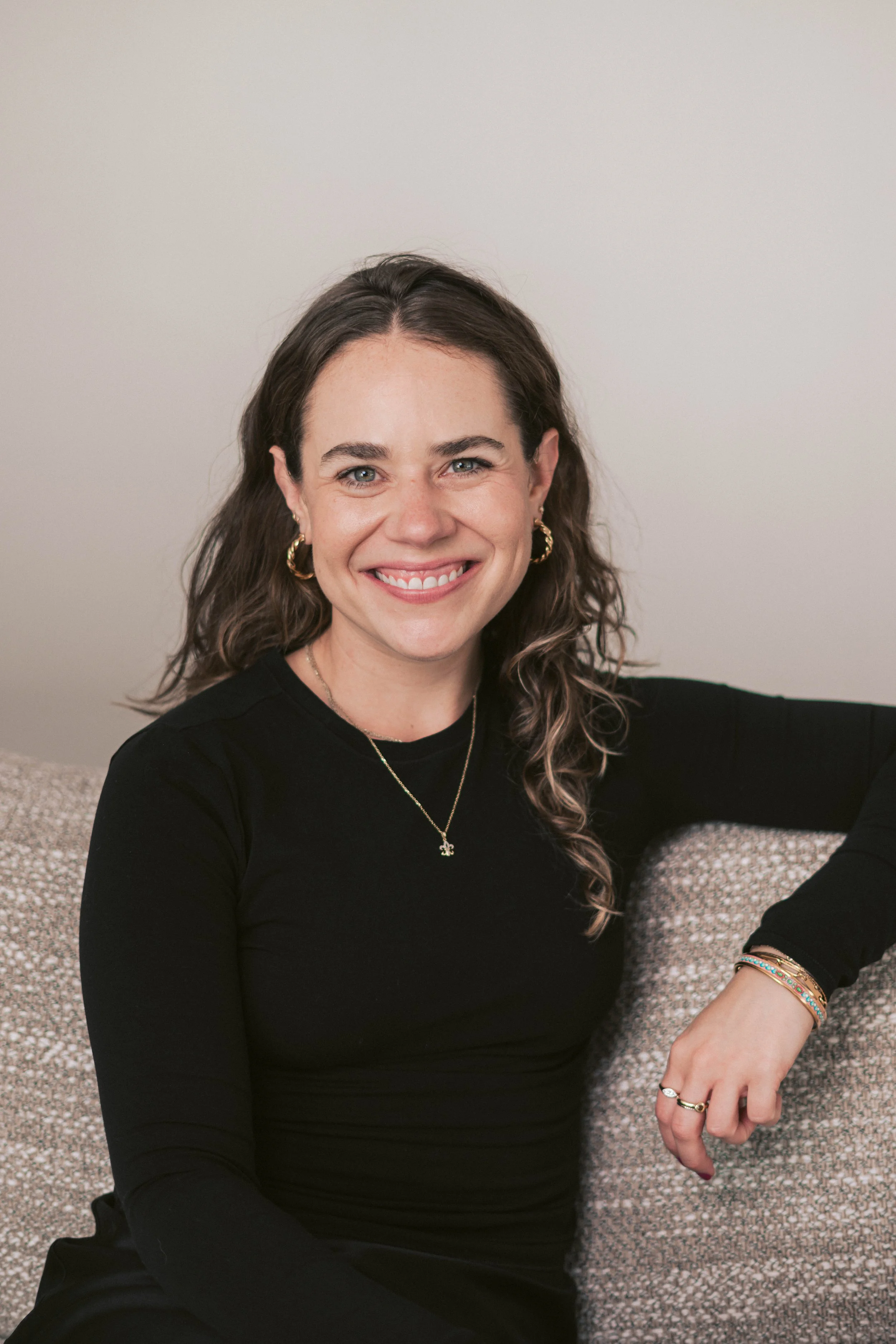 A woman with long, curly brown hair, wearing a black long-sleeve top, gold hoop earrings, a gold necklace, and colorful bracelets, smiling while sitting on a patterned beige couch against a plain light-colored wall.