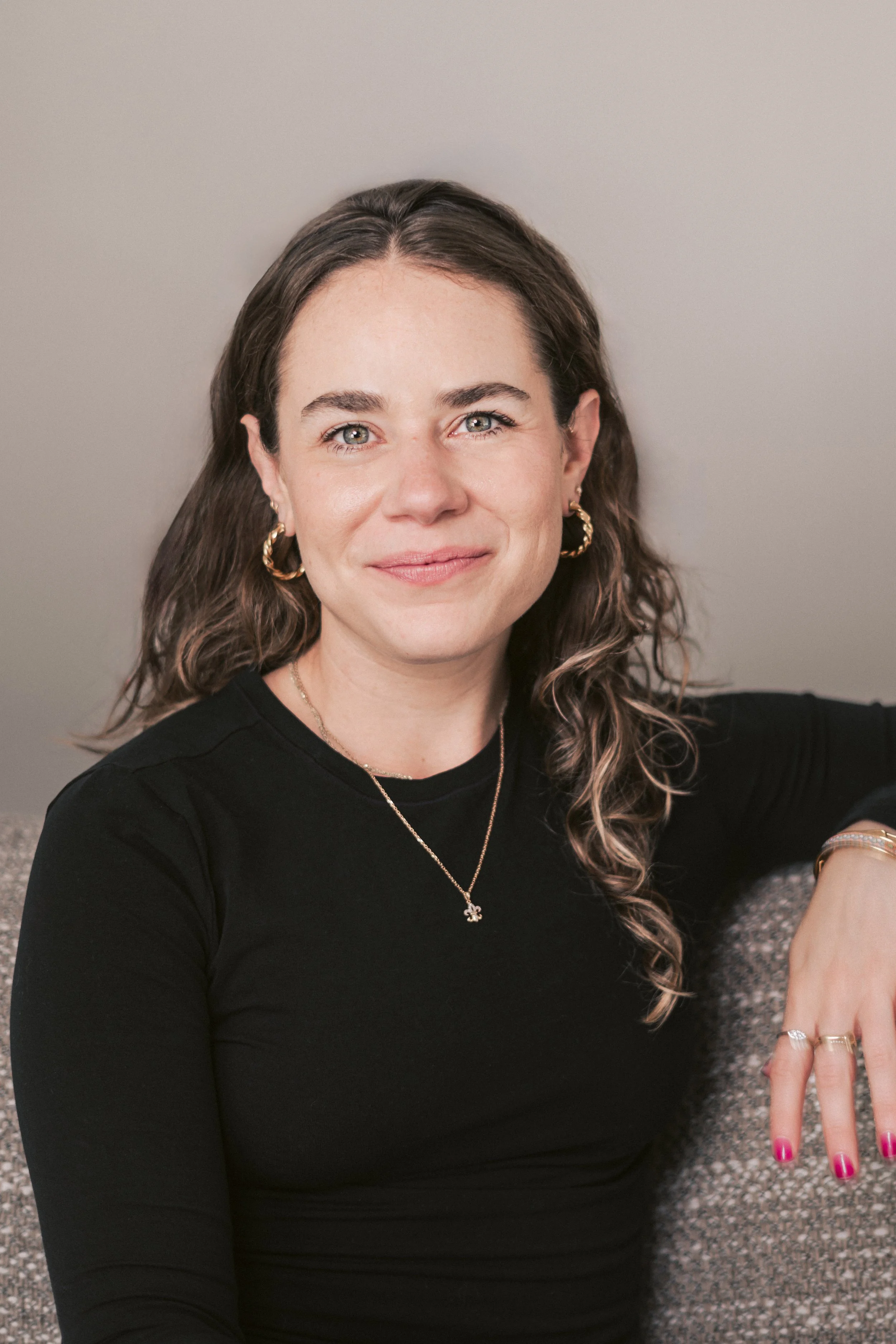 A woman with dark curly hair, wearing a black top, gold hoop earrings, a necklace with a small pendant, and pink nail polish, sitting on a sofa and smiling at the camera.