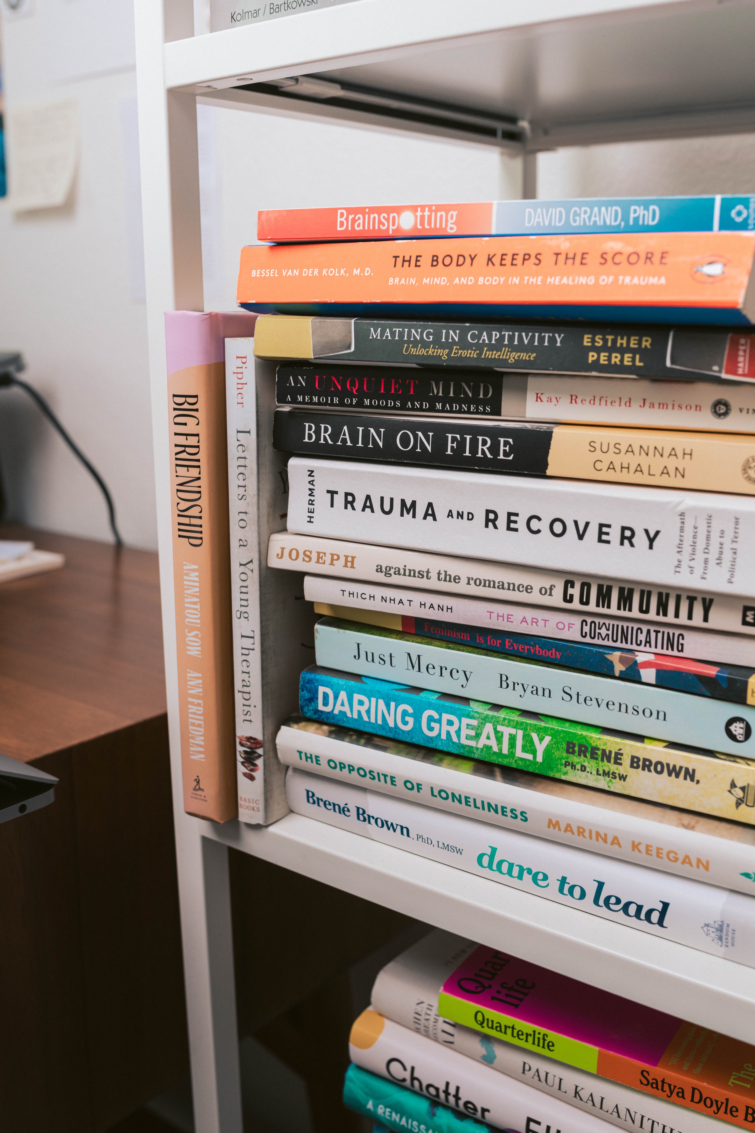 A bookshelf filled with various books on psychology, community, and personal development. Some titles include "Brainspotting" by David Grand, "The Body Keeps the Score," "Big Friendship," "Trauma and Recovery," "Just Mercy," "Daring Greatly," and "Dare to Lead."