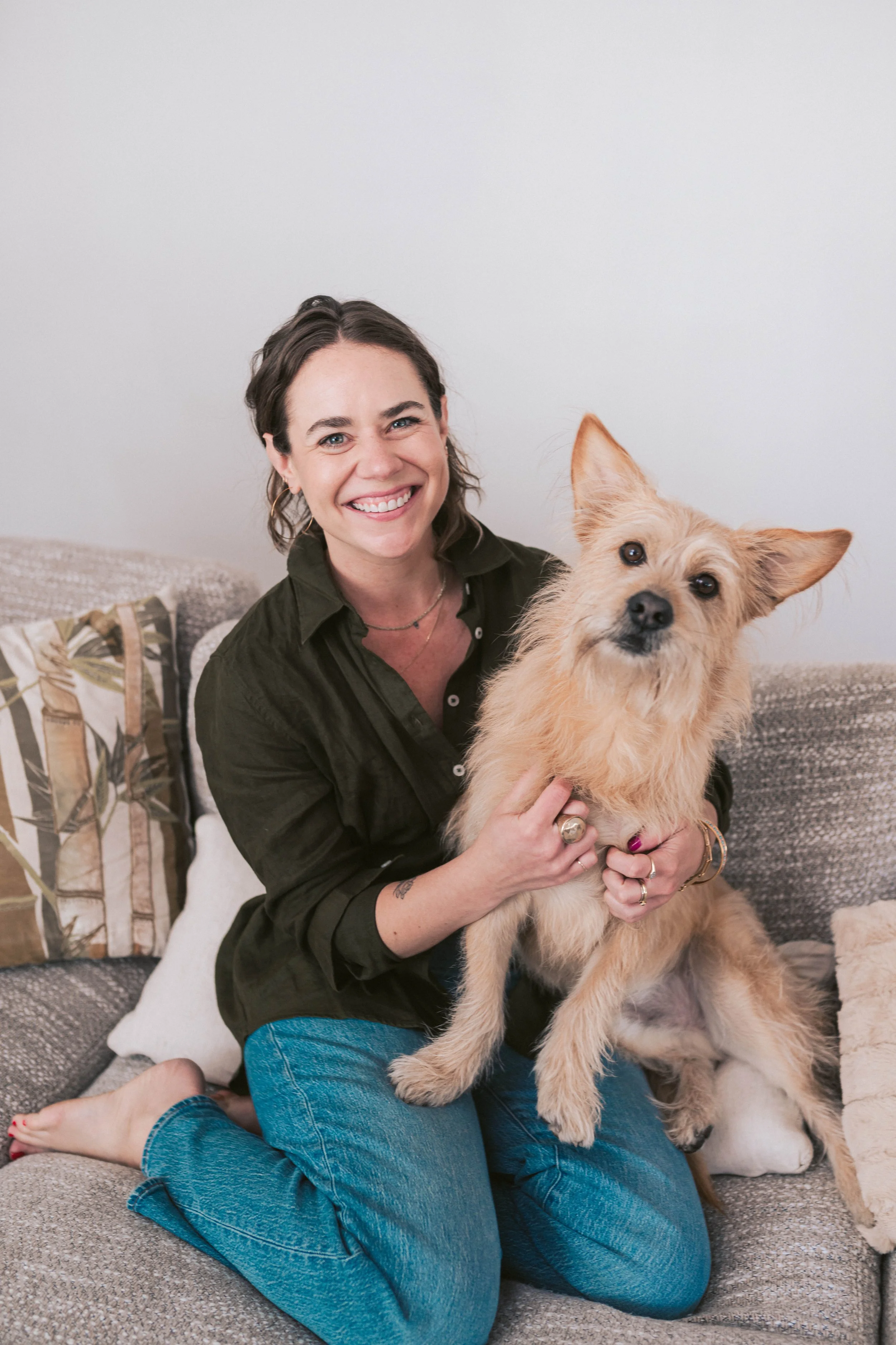 A smiling woman sitting on a couch holding a tan mixed-breed dog, both looking at the camera in a cozy living room.