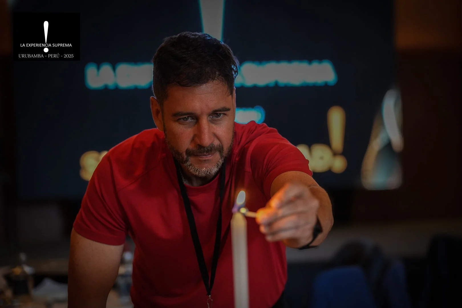 Hombre con barba y camiseta roja, encendiendo una vela con un fósforo en un entorno oscuro.