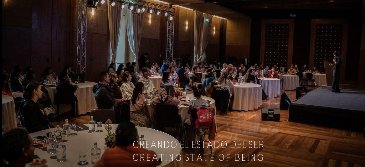 Imagen de un evento o conferencia con muchas personas sentadas en mesas redondas, asistiendo a una presentación en un escenario con una oradora hablando.