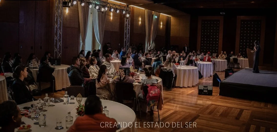 Personas en una conferencia en un auditorio con escenario, algunas tomando notas y otras escuchando, en un ambiente formal con mesas redondas y decoración elegante.