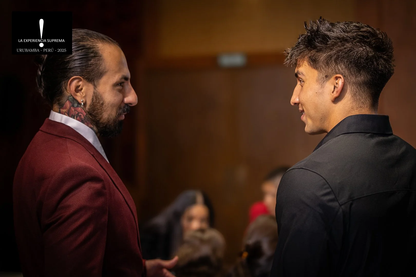 Dos hombres sonriendo y mirándose uno al otro en una conversación en un entorno interior, con fondo de colores oscuros y personas difusas en el fondo.