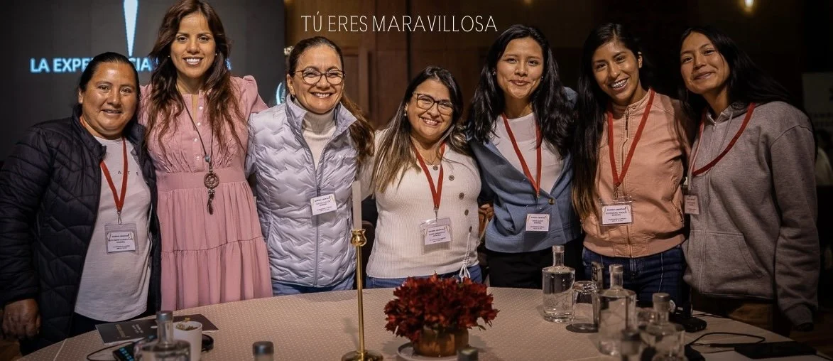 Grupo de siete mujeres sonriendo, de pie detrás de una mesa con jarrones y una flor. Están en un evento, con medallas colgadas al cuello y fondo con letreros.