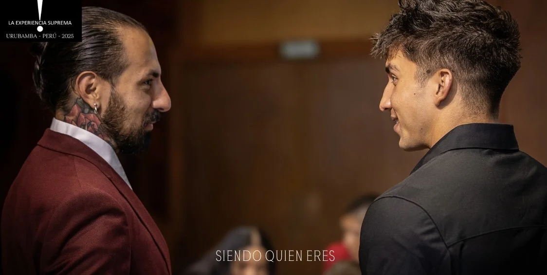 Dos hombres sonriendo y mirándose uno al otro en una conversación en un entorno interior, con fondo de colores oscuros y personas difusas en el fondo.