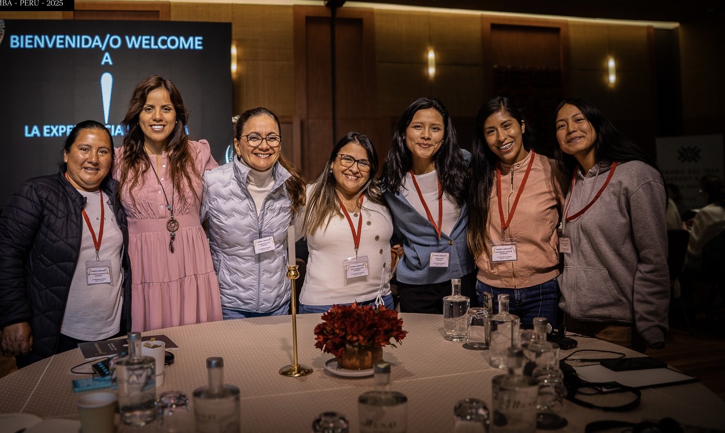 Grupo de siete mujeres sonriendo en una conferencia, con gafetes colgados del cuello, frente a una mesa con varias botellas y un florero, en un ambiente profesional.