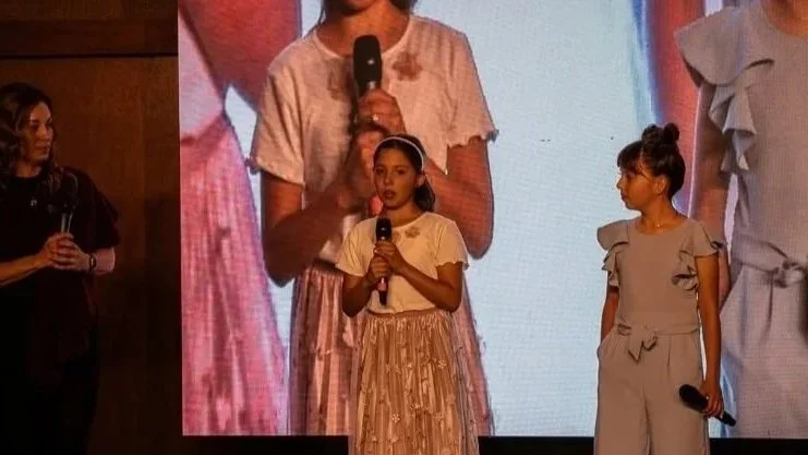 Tres niñas de pie en un escenario con micrófonos y una mujer a la izquierda, posiblemente durante un evento o presentación.