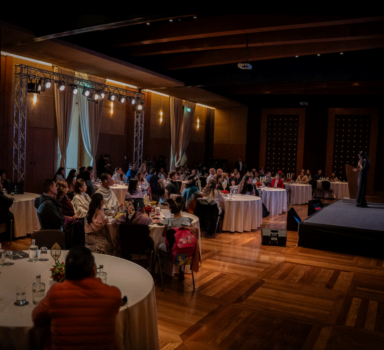 Una conferencia o evento en un salón con espectadores sentados en mesas redondas, escuchando a una oradora en un podio, con iluminación y decoración elegante.