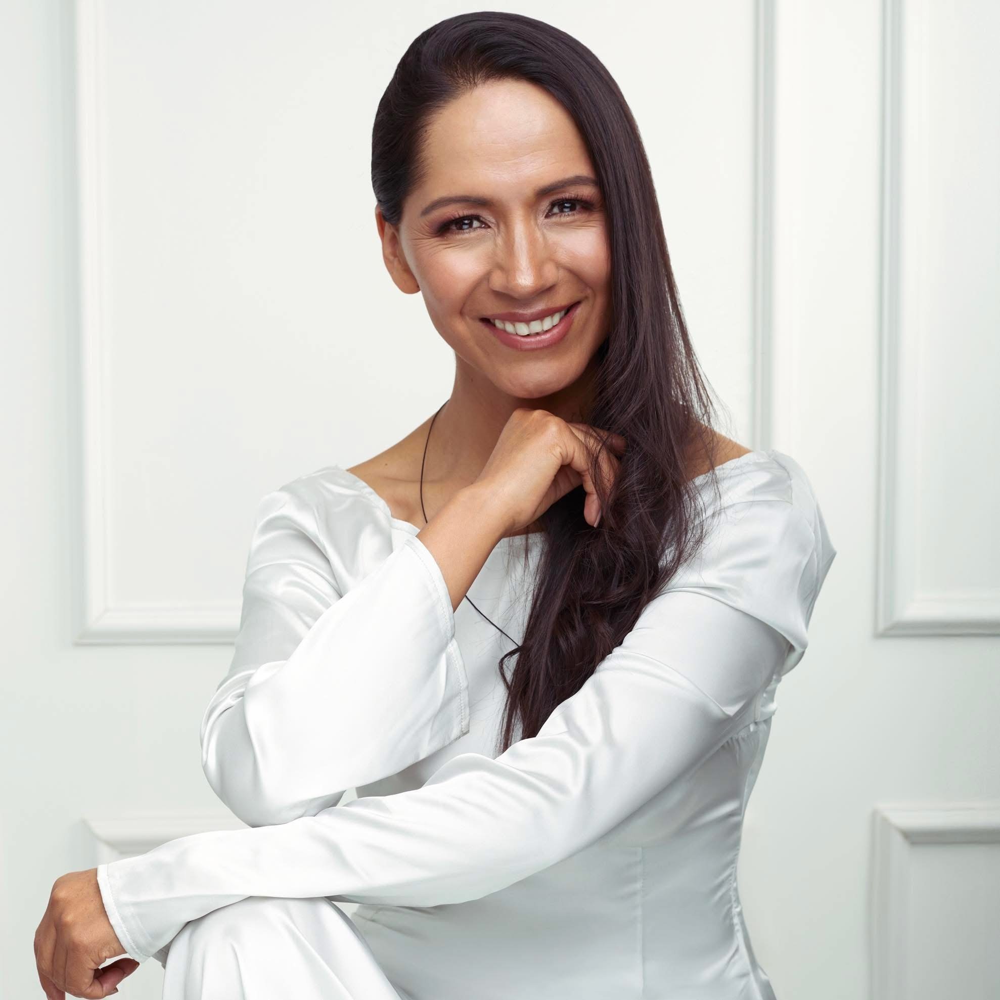 Mujer sonriendo con cabello largo y oscuro, vistiendo blusa blanca, en un entorno interior con fondo blanco y molduras.