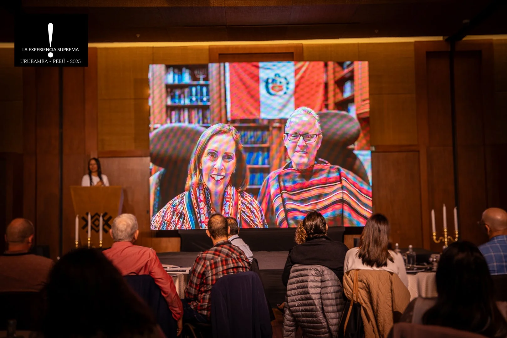 Conferencia o evento formal en un salón amplio con asistentes sentados en mesas redondas y rectas, enfocando a una mujer en el escenario que habla desde un podio.