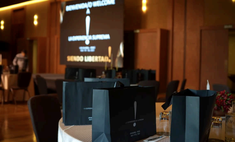 Mesas vestidas con manteles blancos en un salón de eventos con bolsas negras en la mesa y una pantalla grande en el fondo con texto en español.