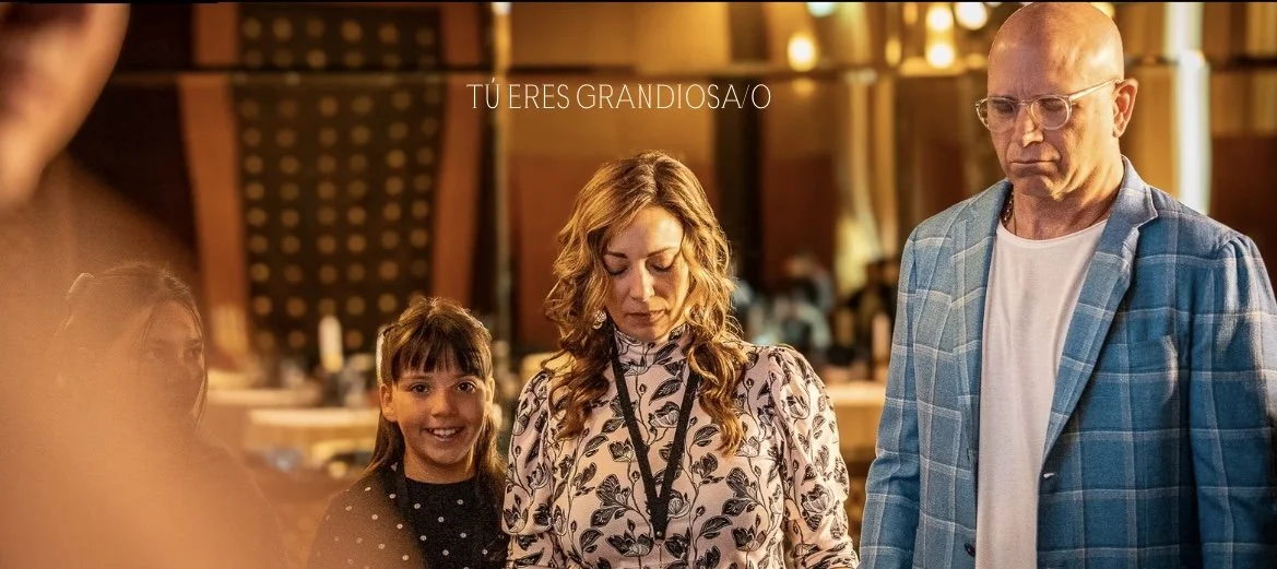 Varias personas en un lugar con ambiente cálido y decorado con madera, una mujer con cabello rizado y ropa con estampado floral, un niño sonriente, un hombre con gafas y camisa con chaqueta a cuadros, y una mujer rubia con expresión pensativa.