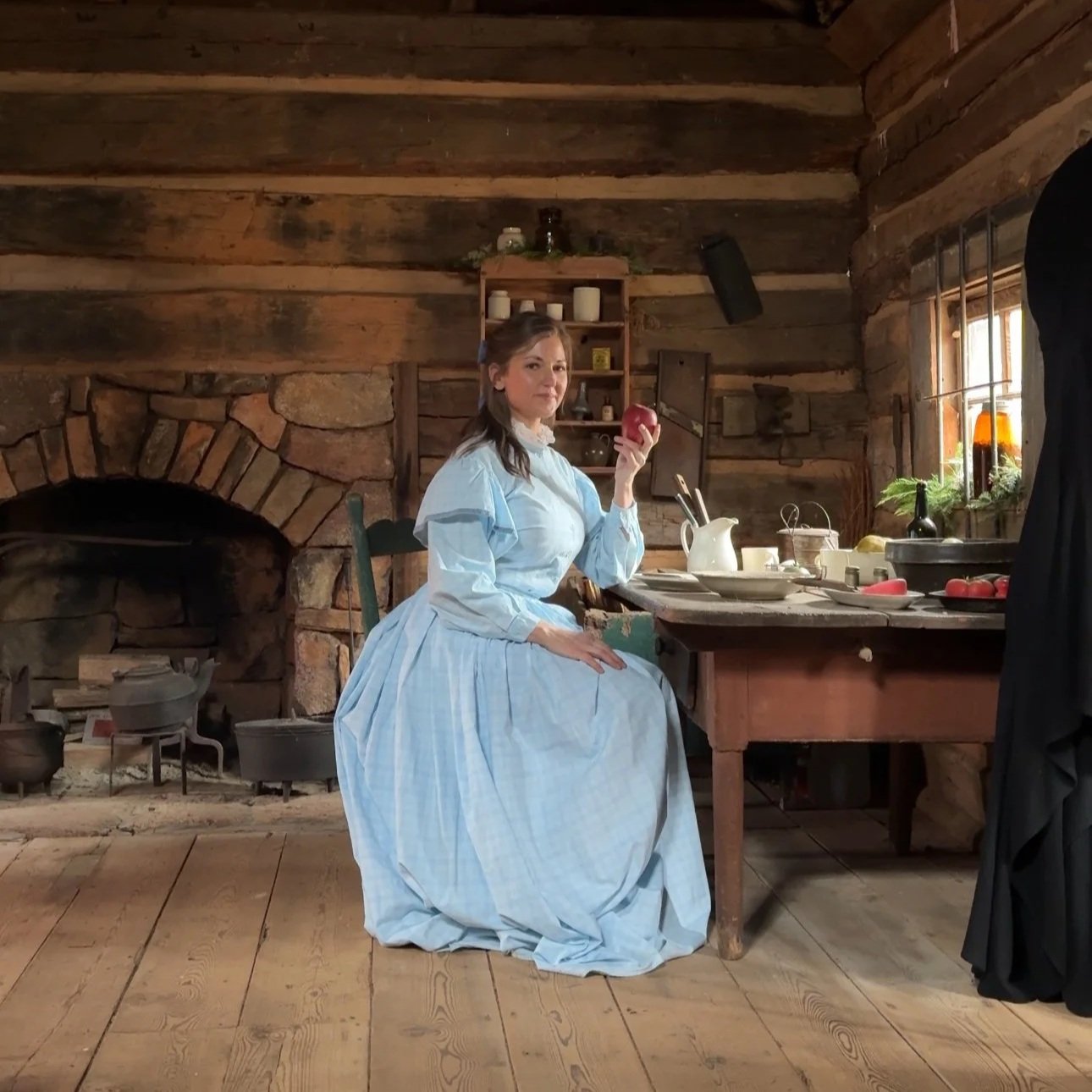 A woman dressed in 19th-century style light blue dress with long sleeves and full skirt, sitting in a rustic kitchen with a stone fireplace and wooden walls, holding a red apple in her hand.