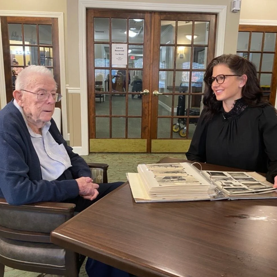 An WW2 Veteran and a woman wearing glasses sitting across from each other at a wooden table, looking at a photo album in a room with glass doors