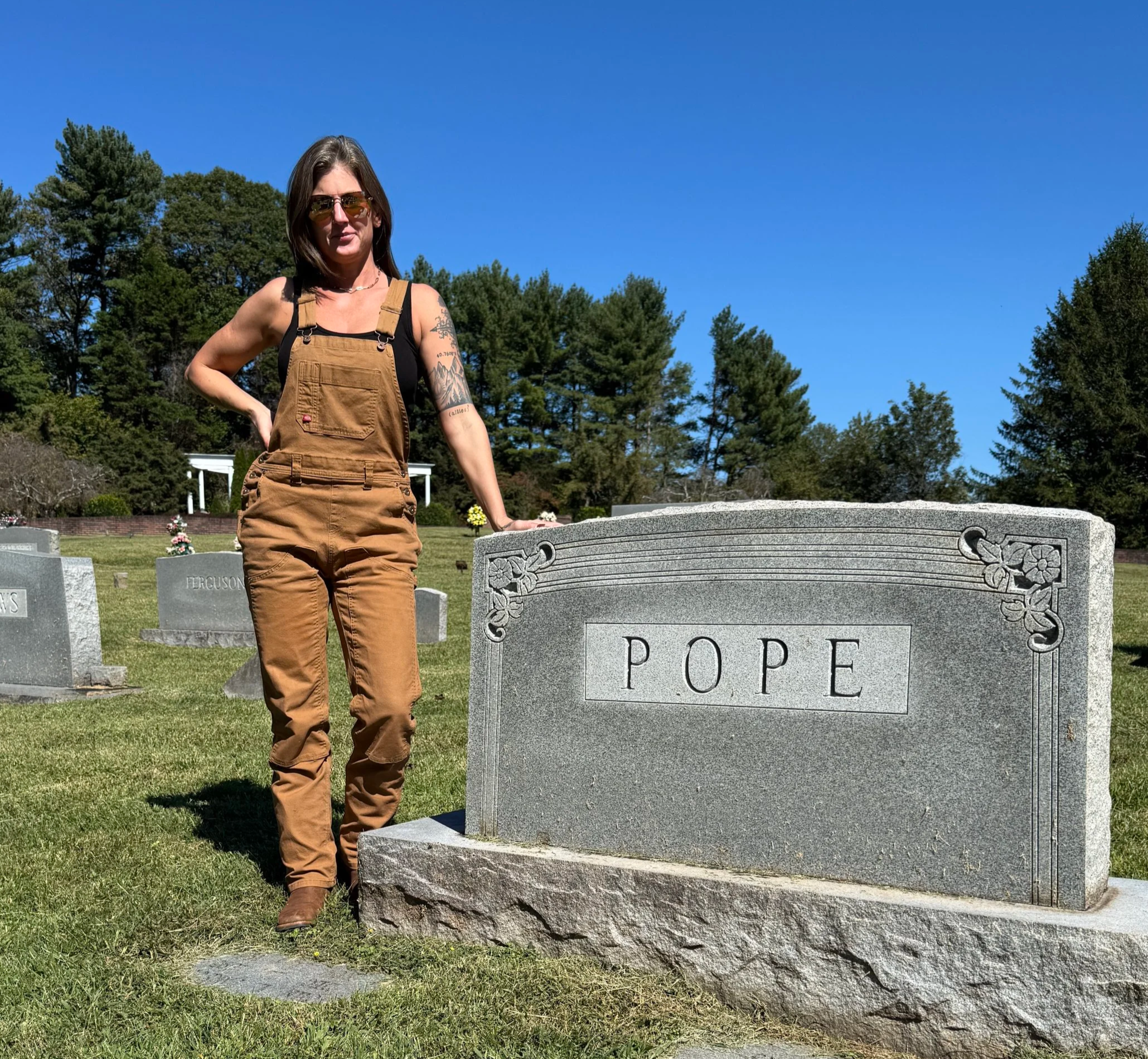 A woman in brown overalls and sunglasses standing next to a gravestone labeled 'Pope' in a cemetery under a clear blue sky.