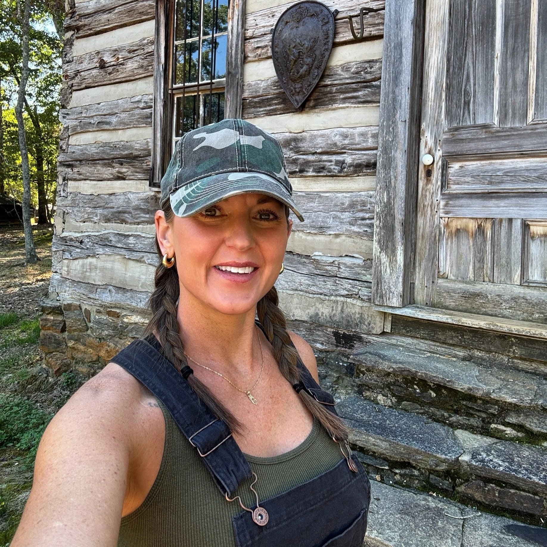 A smiling woman with braided hair, wearing a camouflage cap, earrings, a gold necklace, and black overalls, stands in front of a rustic wooden cabin with window and decorative shield.