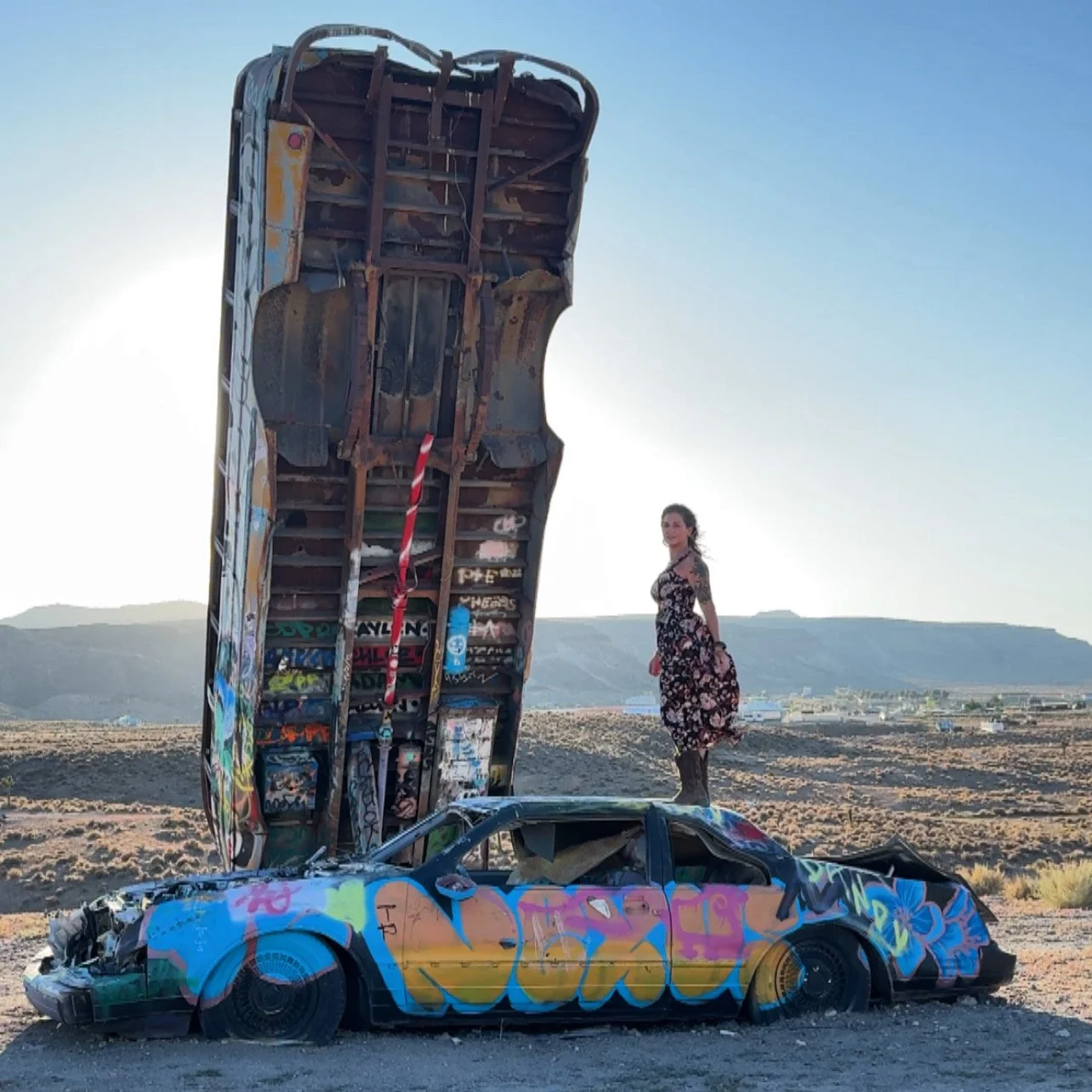A woman standing on top of a graffiti-covered wrecked car with a graffitied, upside-down car hood propped up behind her in a desert landscape.