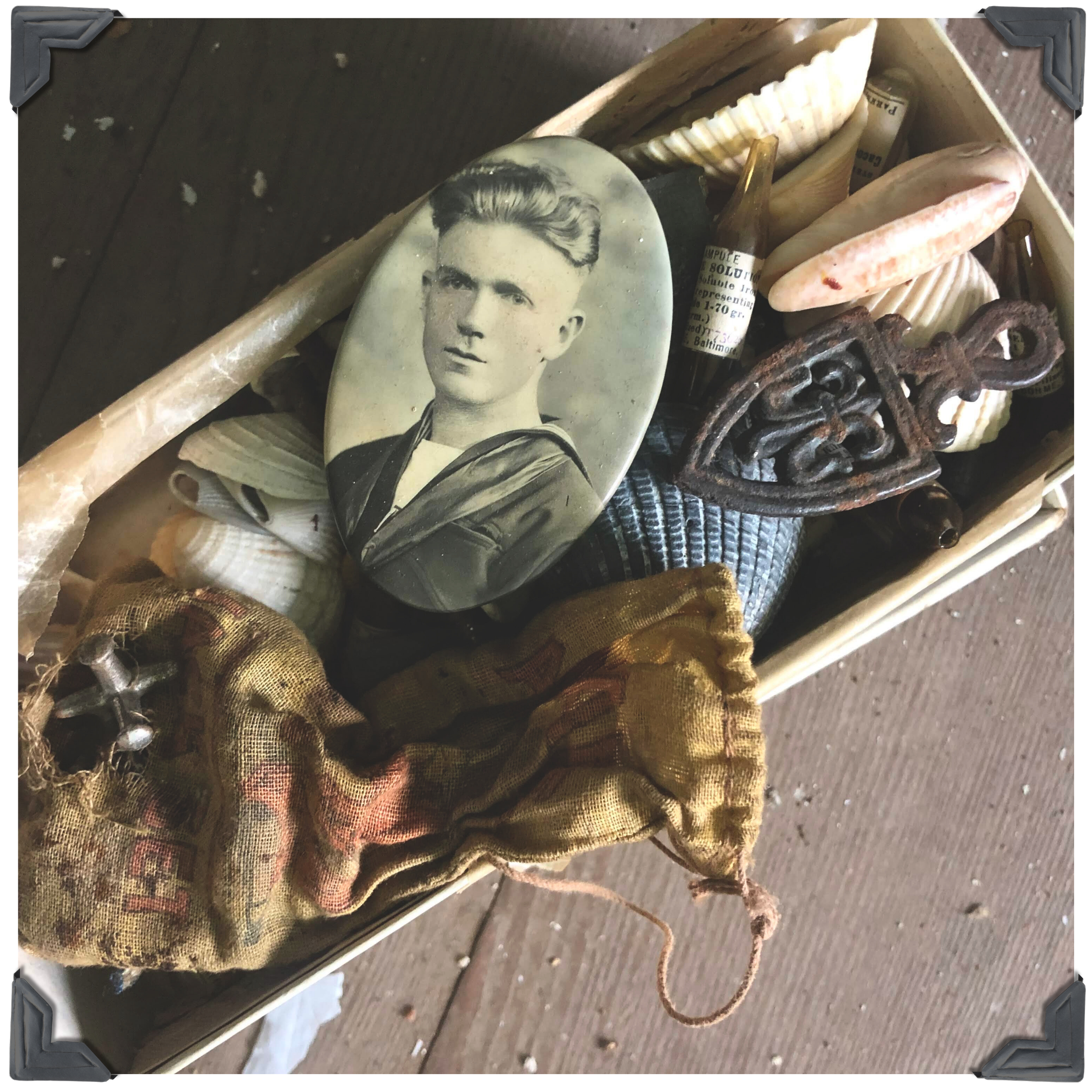 a box of keepsakes lying ont he dusty floor of an old house, that include a worn bag of jacks, an old photo of a sailer, small bottles of medicine, and some sea shells