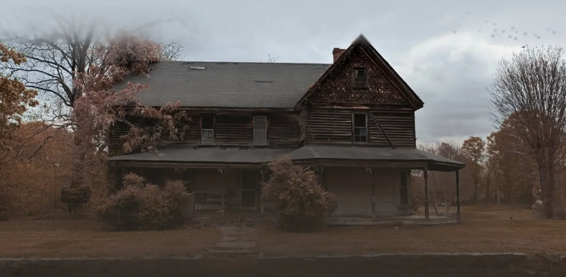 Old, abandoned wooden house with broken windows and a sagging porch, surrounded by leafless trees and bushes, under a cloudy sky.