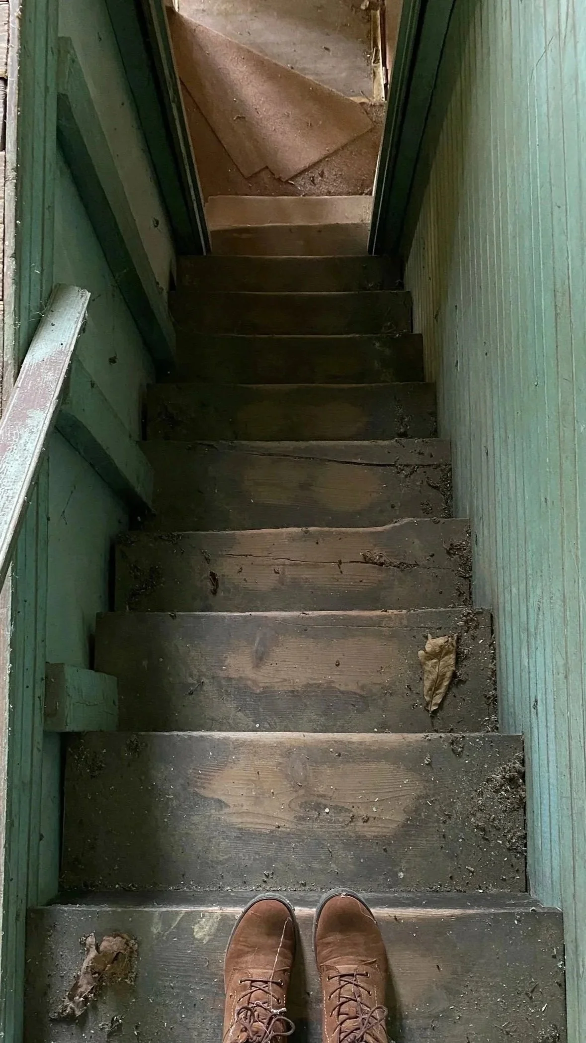 A person's feet wearing brown boots stand on a wooden staircase leading down to a dirt and concrete surface outside. The staircase is old and weathered, with some dirt and debris on the steps.
