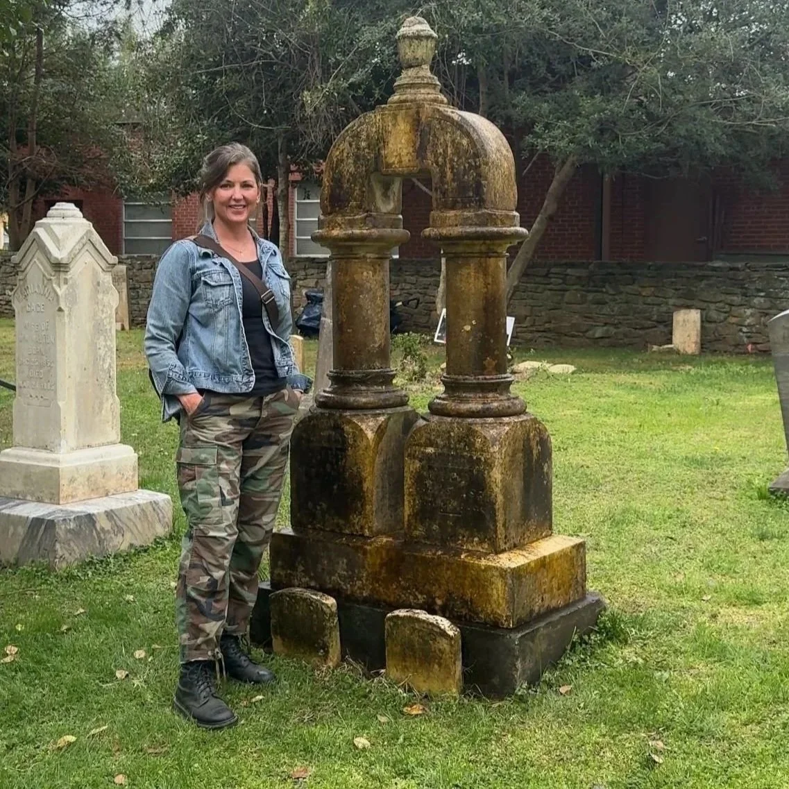 Woman with denim jacket and camouflage pants standing next to an old, moss-covered headstone in a cemetery.