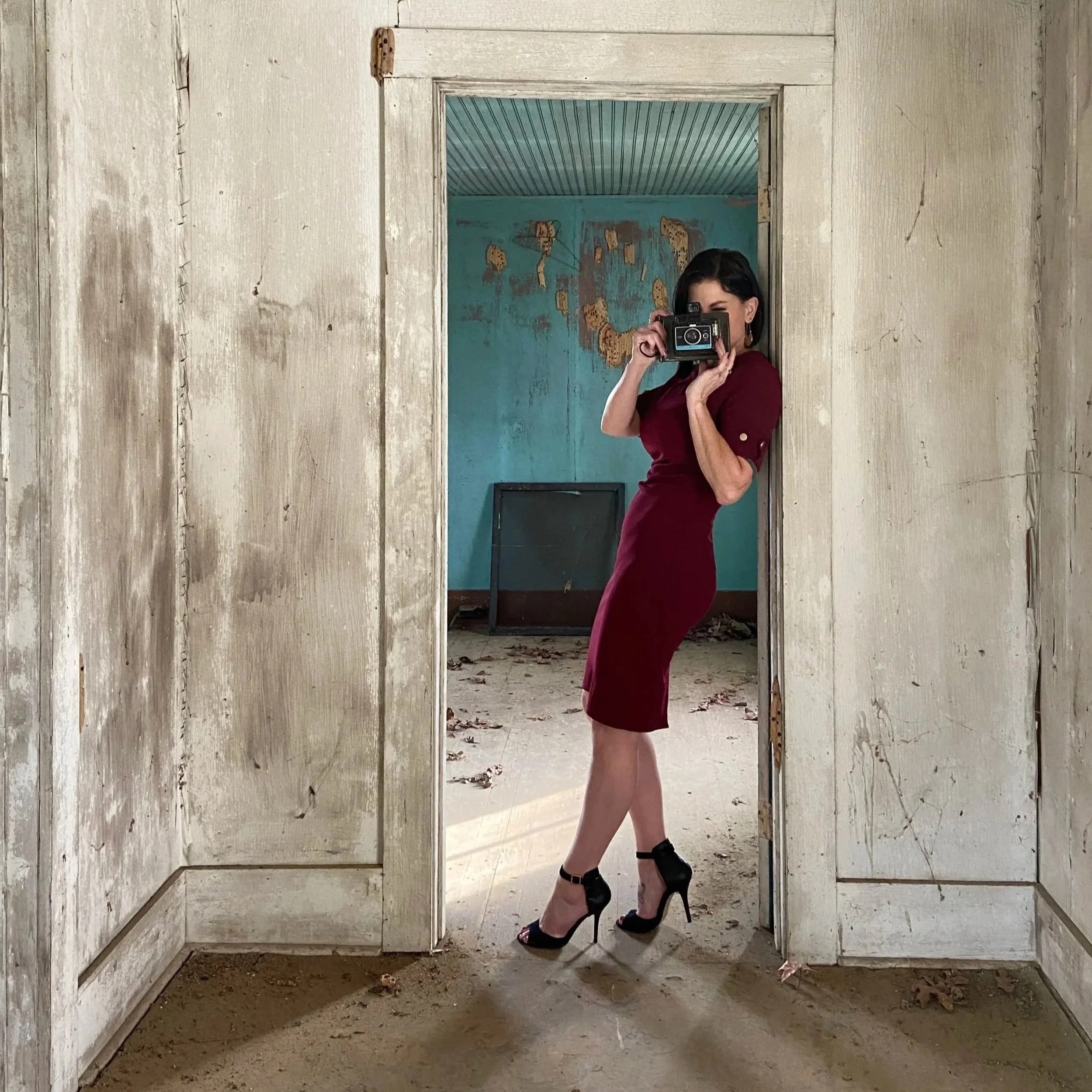 A woman taking a selfie with a vintage camera in an empty, dilapidated room with peeling paint and dirt on the floor, wearing a fitted burgundy dress and black high-heeled shoes.