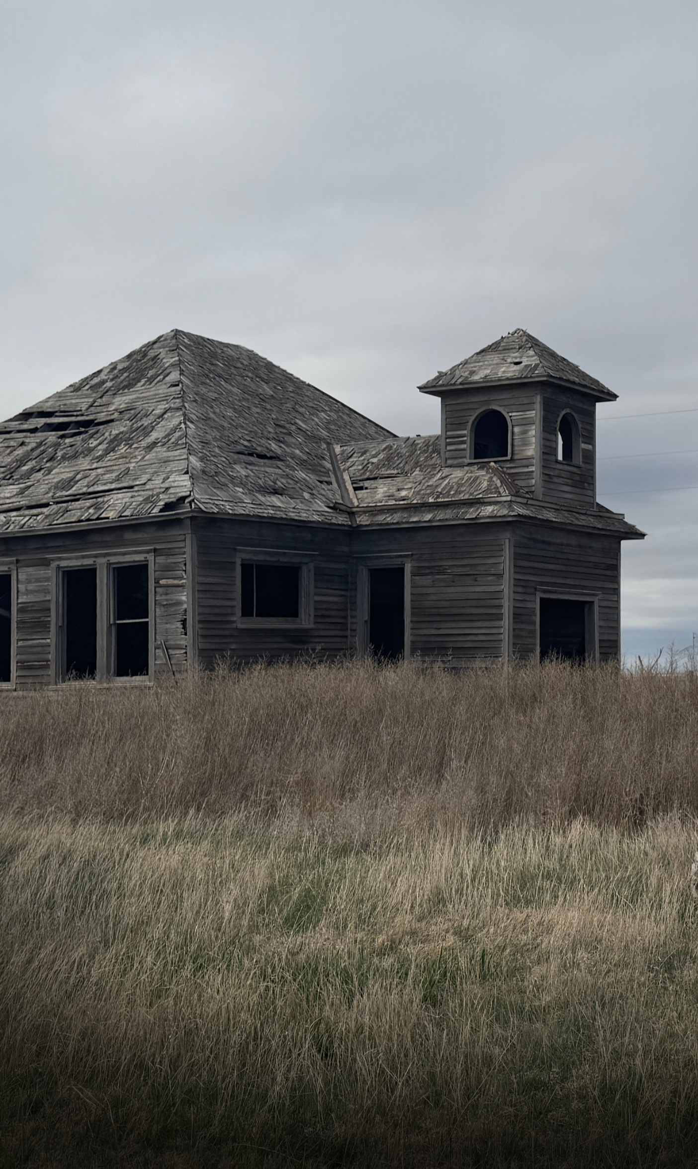An abandoned, dilapidated wooden house with broken windows and a deteriorated roof, surrounded by overgrown grass under a cloudy sky.