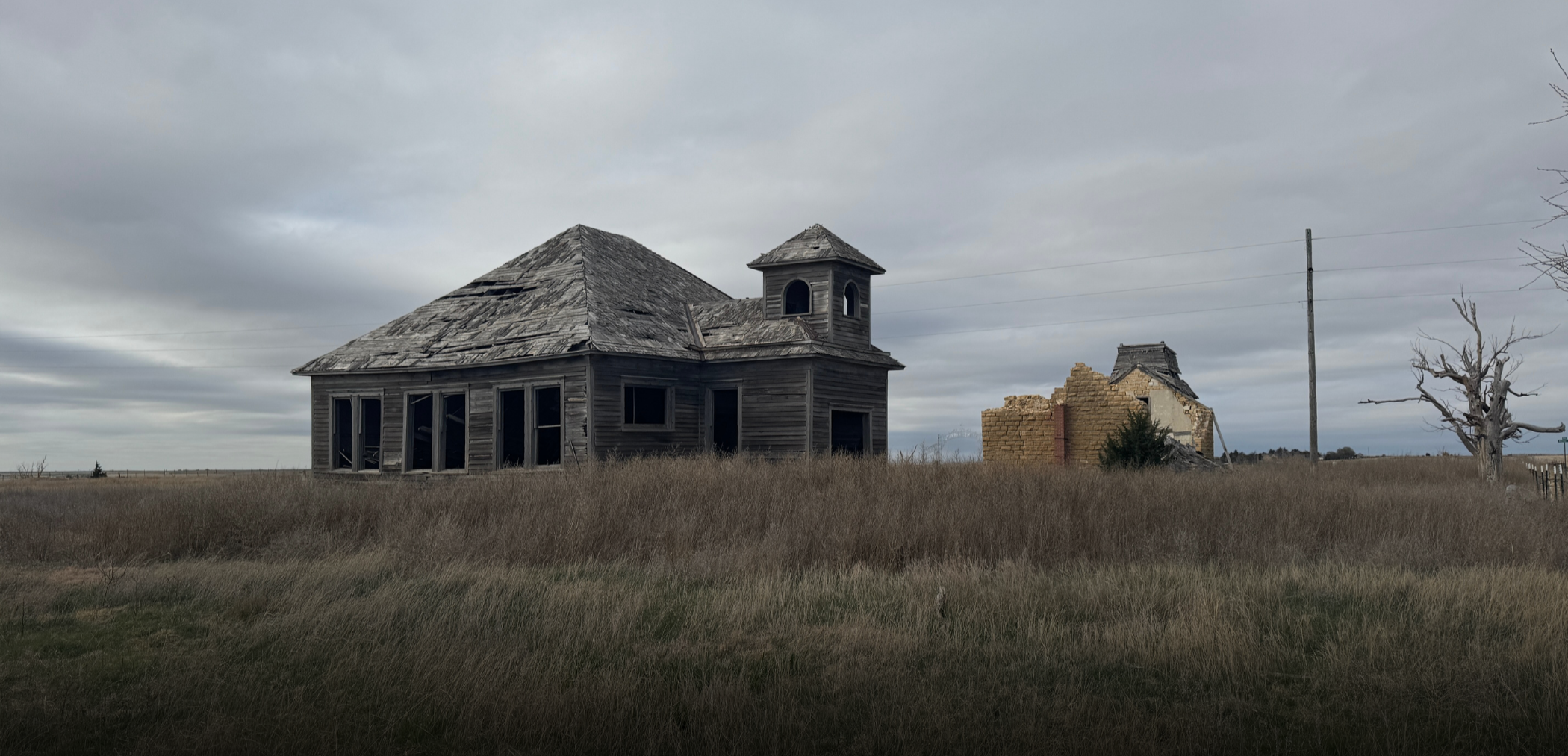Old, abandoned wooden house with broken windows and a damaged roof in a grassy field under a cloudy sky. There is also a partially collapsed brick building, a utility pole, and a leafless tree nearby.