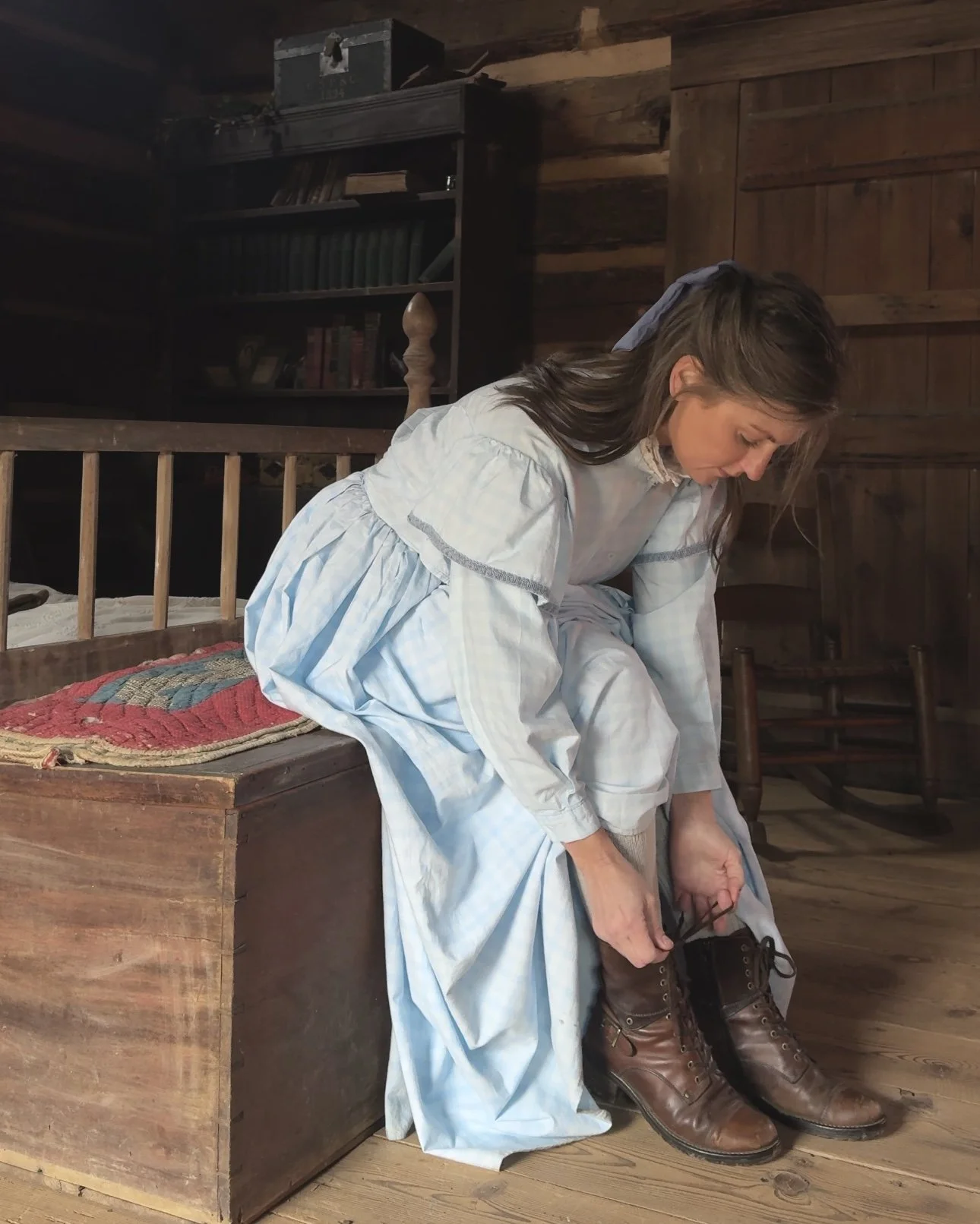 A woman in vintage clothing tying her leather boots inside a rustic wooden room.