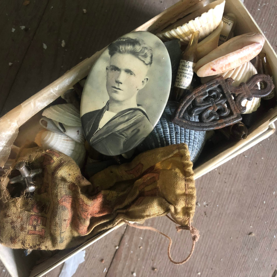 a box of keepsakes lying on the dusty floor of an old house, that include a worn bag of jacks, an old photo of a sailer, small bottles of medicine, and some sea shells