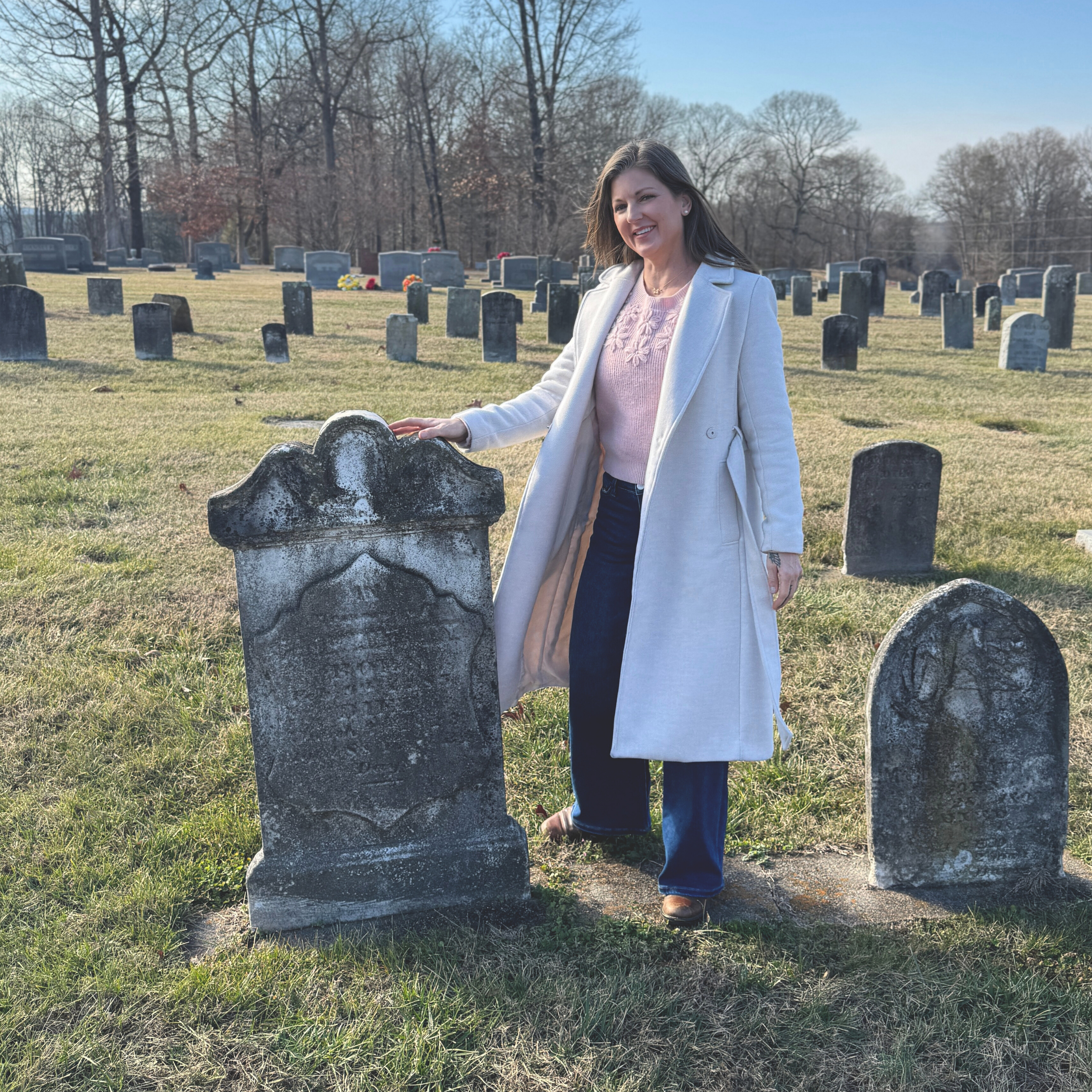 Woman in a white coat standing next to a gravestone in a cemetery on a clear day.