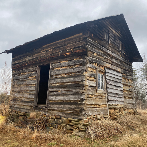 Old worn-down wooden log cabin on a stone foundation with a missing front door and small window, surrounded by dry grass and leafless trees under a cloudy sky.