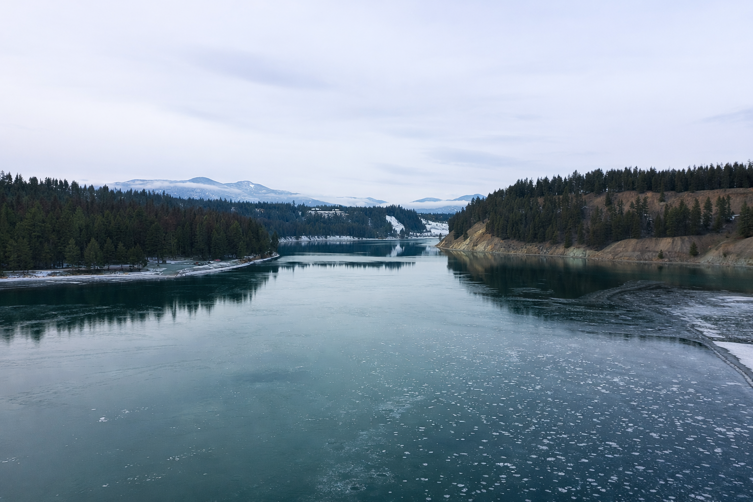 A wide river surrounded by forested hills and distant snow-capped mountains under a cloudy sky.