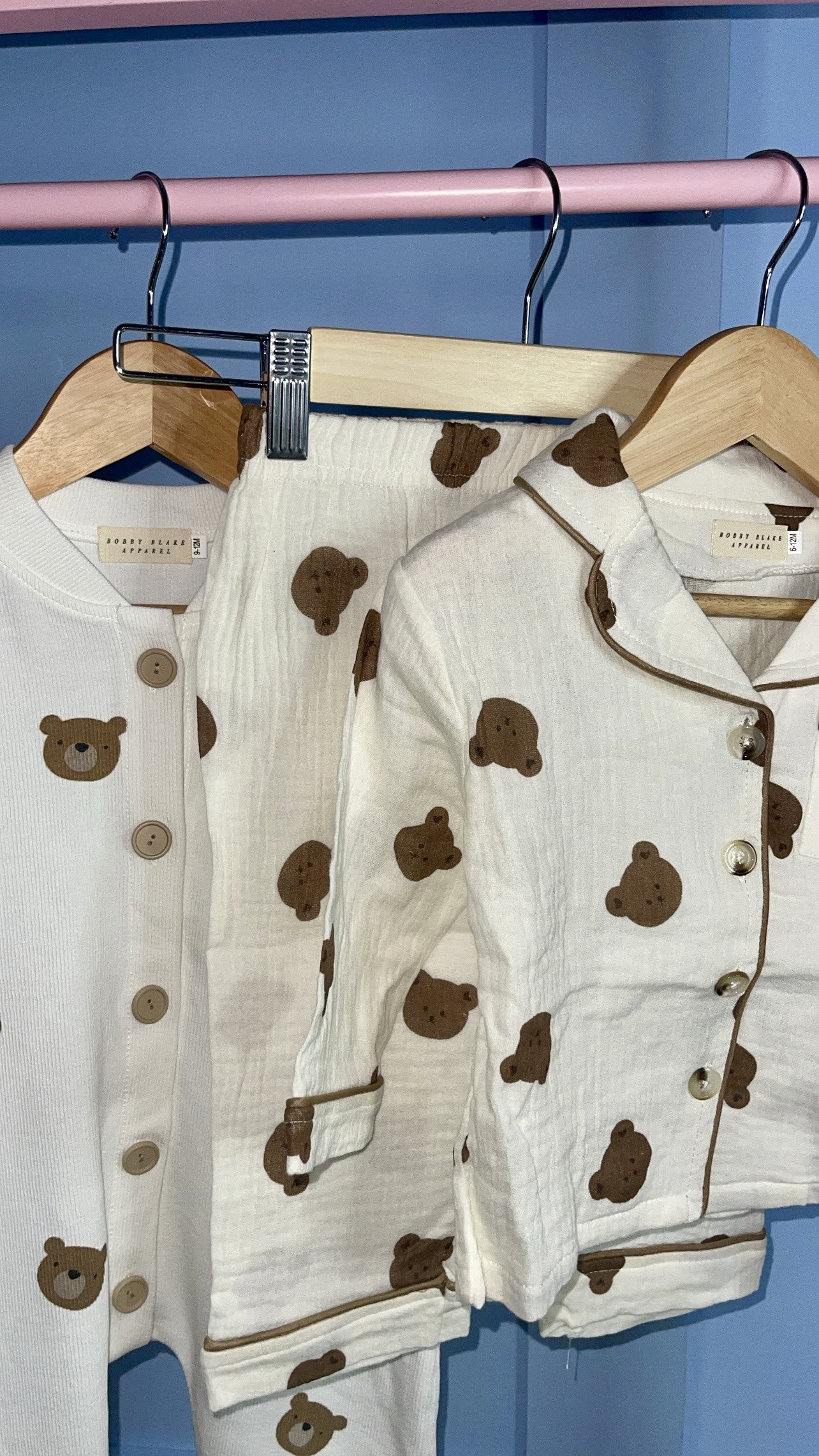 Three children's pyjamas with teddy bear print hanging on wooden hangers in a store, with a blue background. BOBBY BLAKE APPAREL