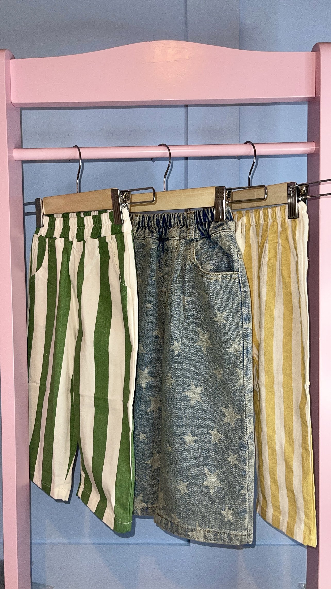 Three pairs of striped and star-patterned pants hanging on a pink and blue clothing rack. BOBBY BLAKE APPAREL