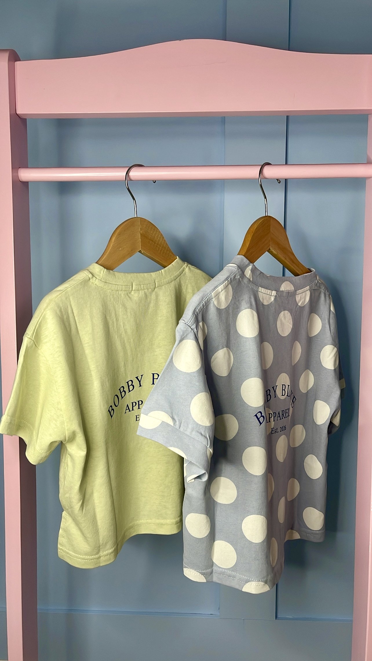 Two T-shirts hanging on wooden hangers inside a pink and blue clothing rack. One yellow T-shirt and one blue T-shirt with large white polka dots, both with the written text 'Bobby Blake Apparel Est. 2026'.